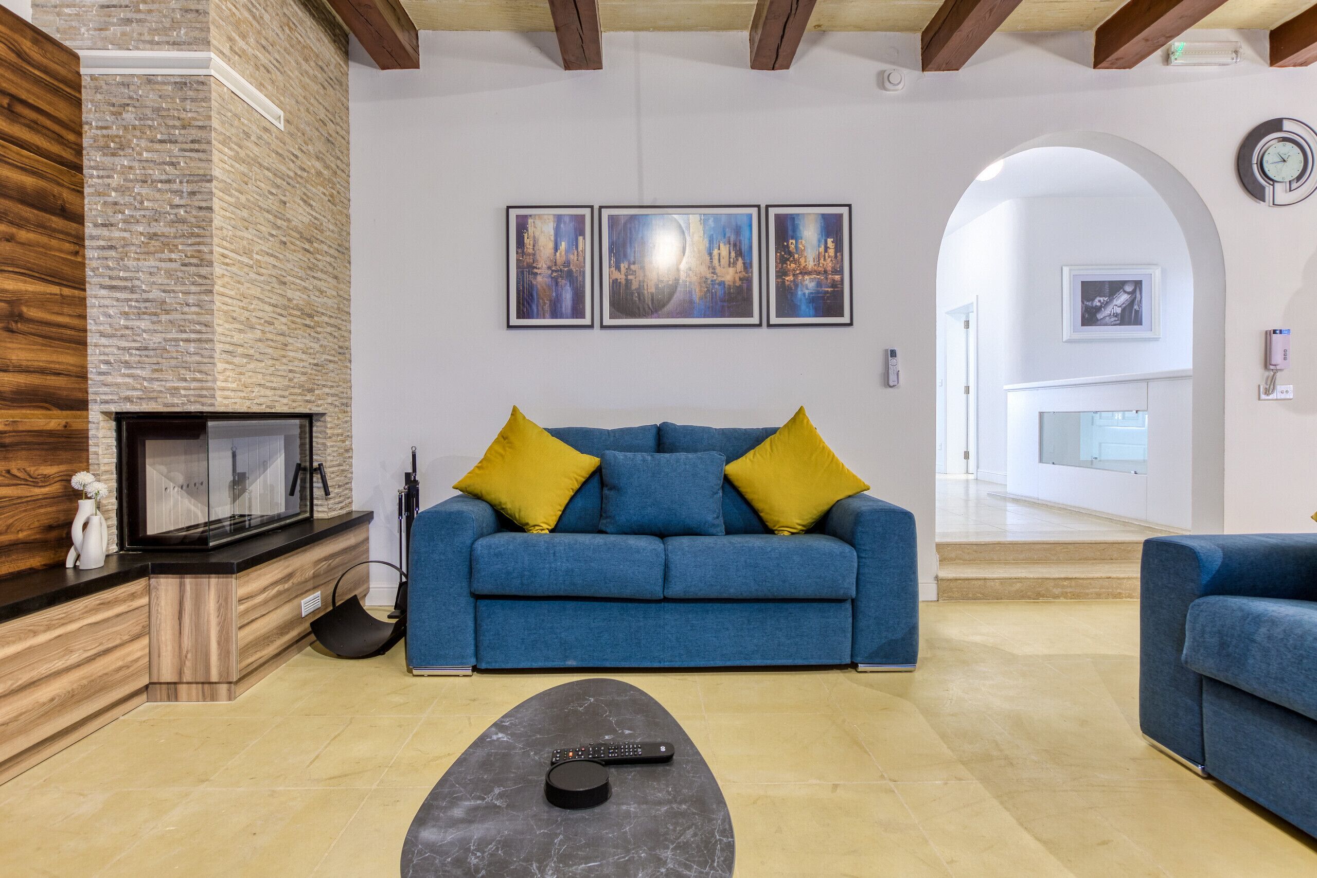 A living room with a blue sofa, yellow cushions, a black coffee table, and a fireplace. There are three framed artworks on the wall and a clock on the wall.