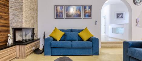 A living room with a blue sofa, yellow cushions, a black coffee table, and a fireplace. There are three framed artworks on the wall and a clock on the wall.