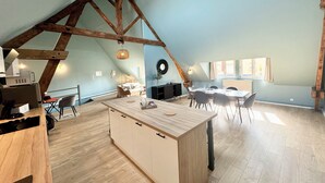 Un loft espacioso con techo alto, vigas de madera a la vista, una isla central con acabados en madera clara y una zona de comedor con mesa y sillas blancas.