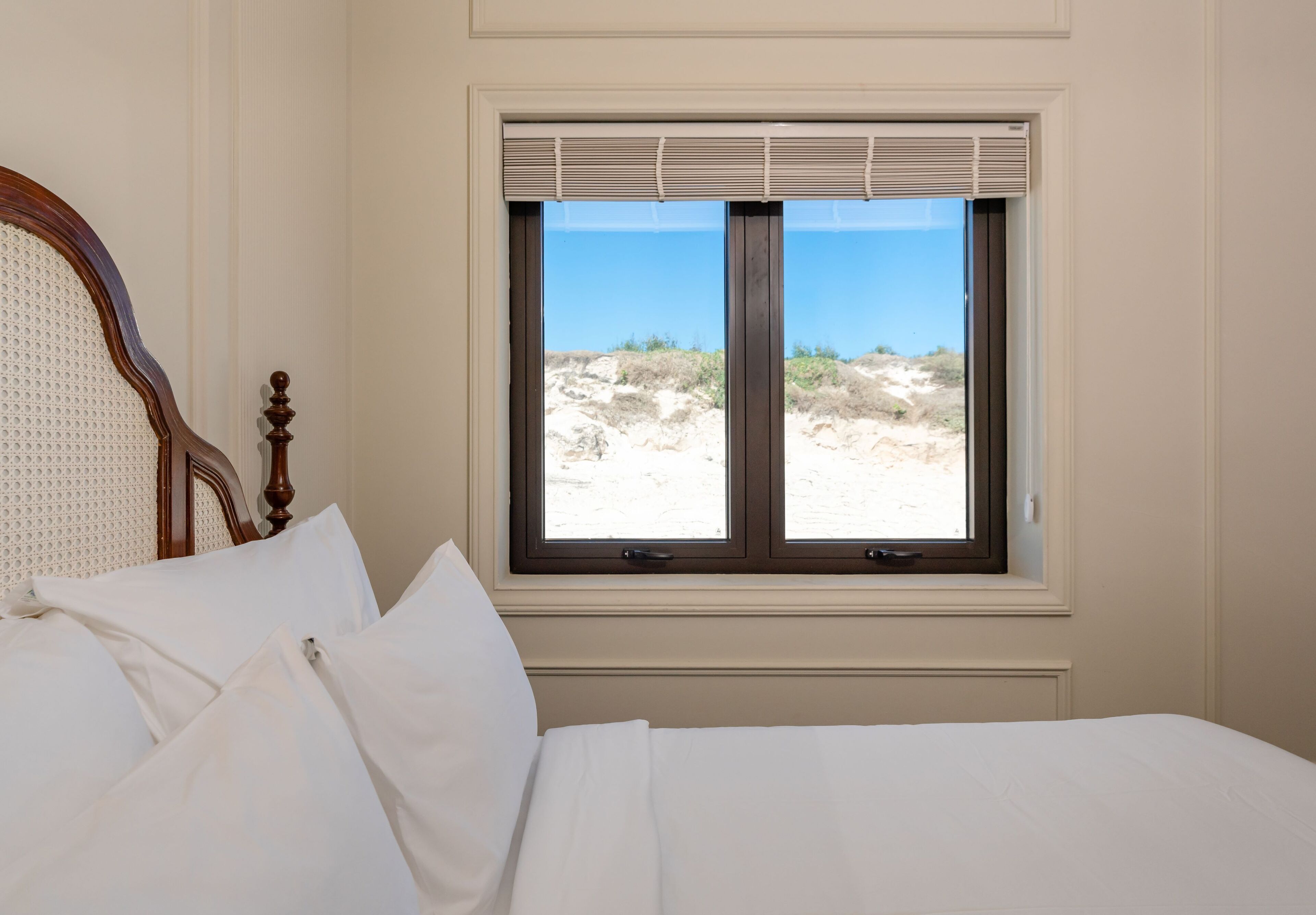 Un lit avec du linge de lit blanc et une tête de lit en bois, une fenêtre avec des stores et une vue sur une plage de sable avec des dunes.