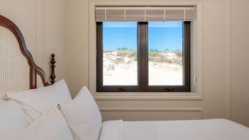 Un lit avec du linge de lit blanc et une tête de lit en bois, une fenêtre avec des stores et une vue sur une plage de sable avec des dunes.
