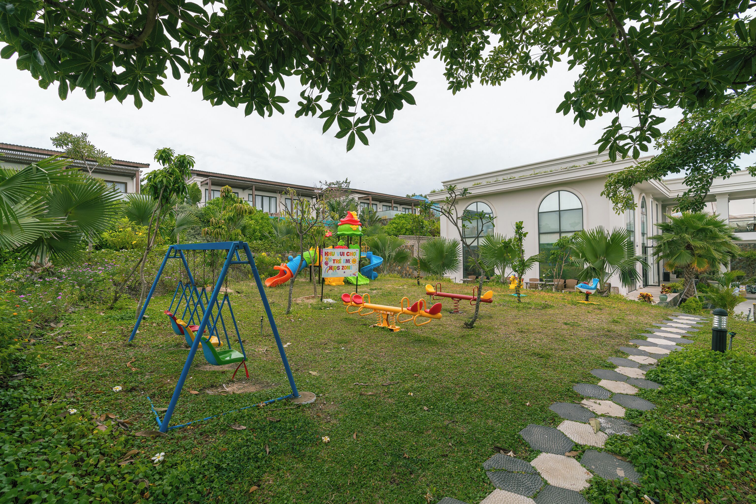 Un parque infantil con columpios y balancines, rodeado de césped y árboles, con un edificio al fondo.
