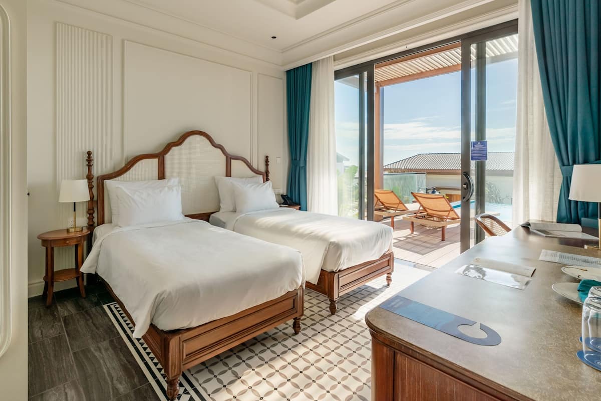 A hotel room with two beds, a balcony with chairs, and a desk with a phone and papers.