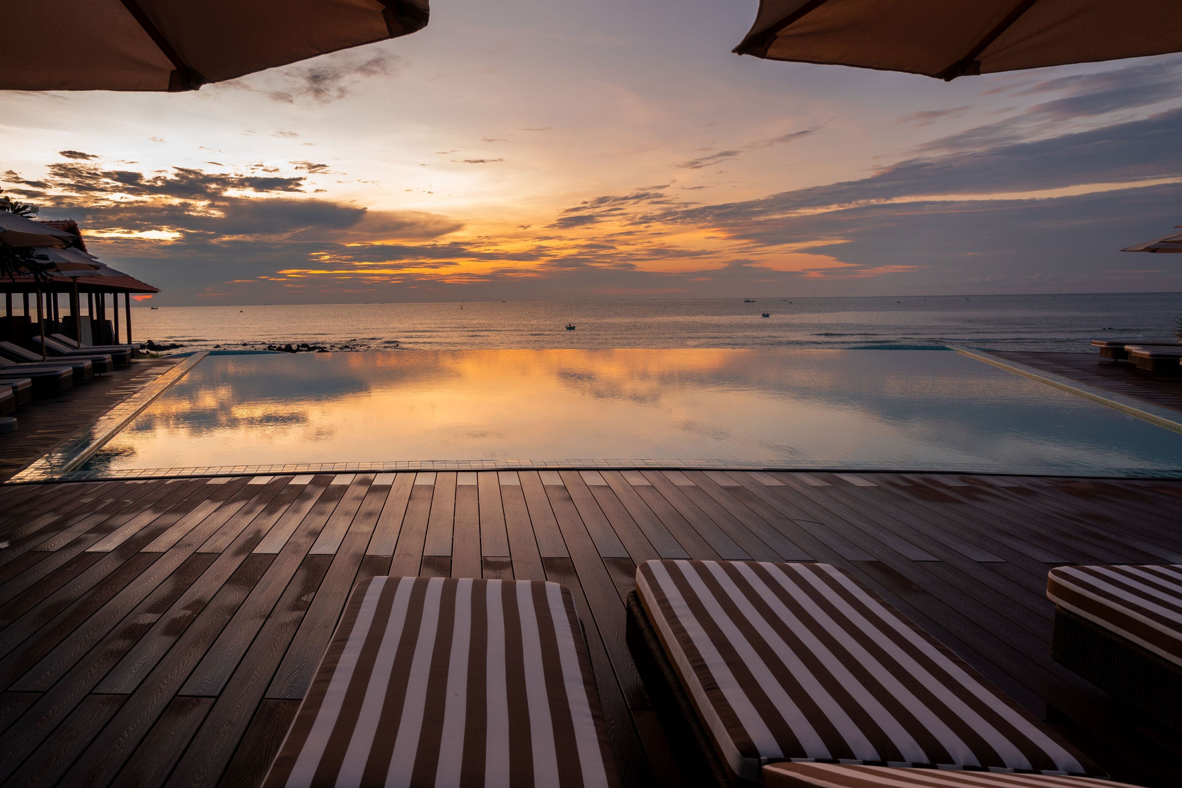 Un terrazzo in legno con poltrone da relax e ombrelloni che si affaccia su una piscina tranquilla e l'oceano al tramonto.