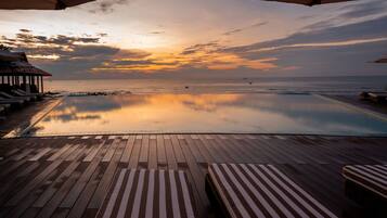 Un terrazzo in legno con poltrone da relax e ombrelloni che si affaccia su una piscina tranquilla e l'oceano al tramonto.