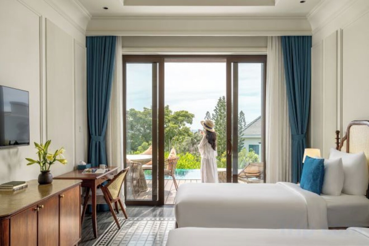 Habitación de hotel con cama, escritorio, televisión y vistas a un balcón con una persona parada.
