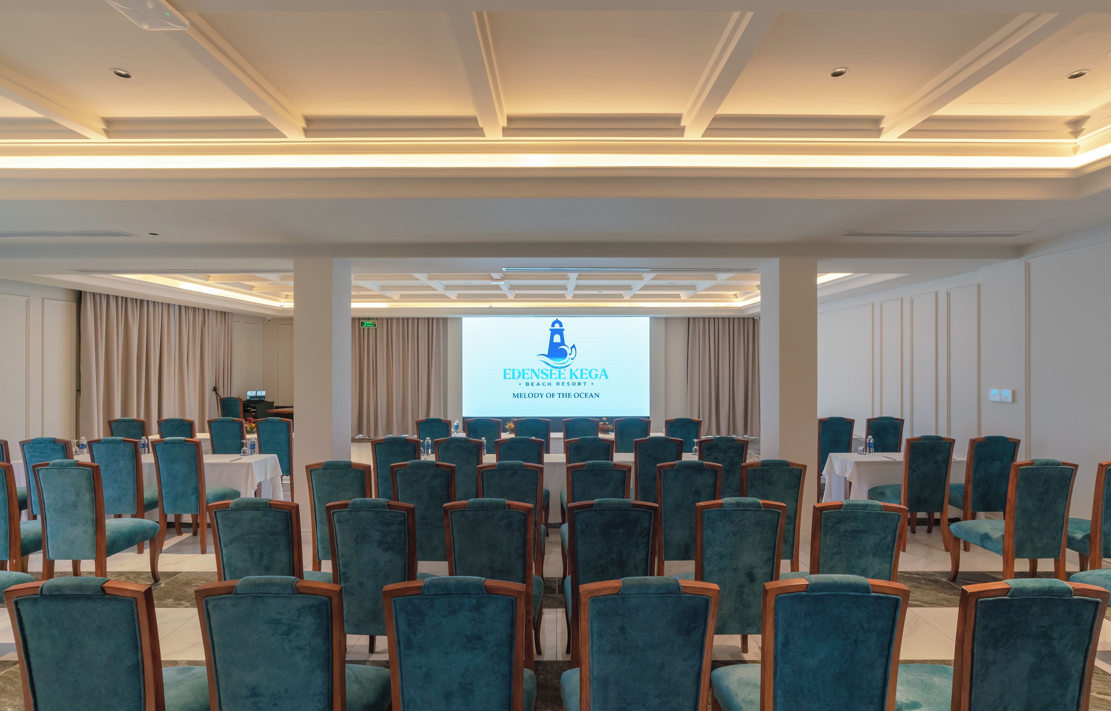 Un salón de conferencias con filas de sillas color turquesa, una pantalla de proyector mostrando 'EDENSEE KEGA BEACH RESORT - MELODY OF THE OCEAN', y un escenario con un atril.