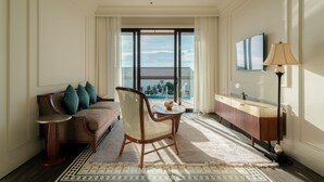 Ein Hotelzimmer mit einer Couch, einem Sessel, einem kleinen Tisch, einer Lampe, einem Fernseher und Blick ins Freie.