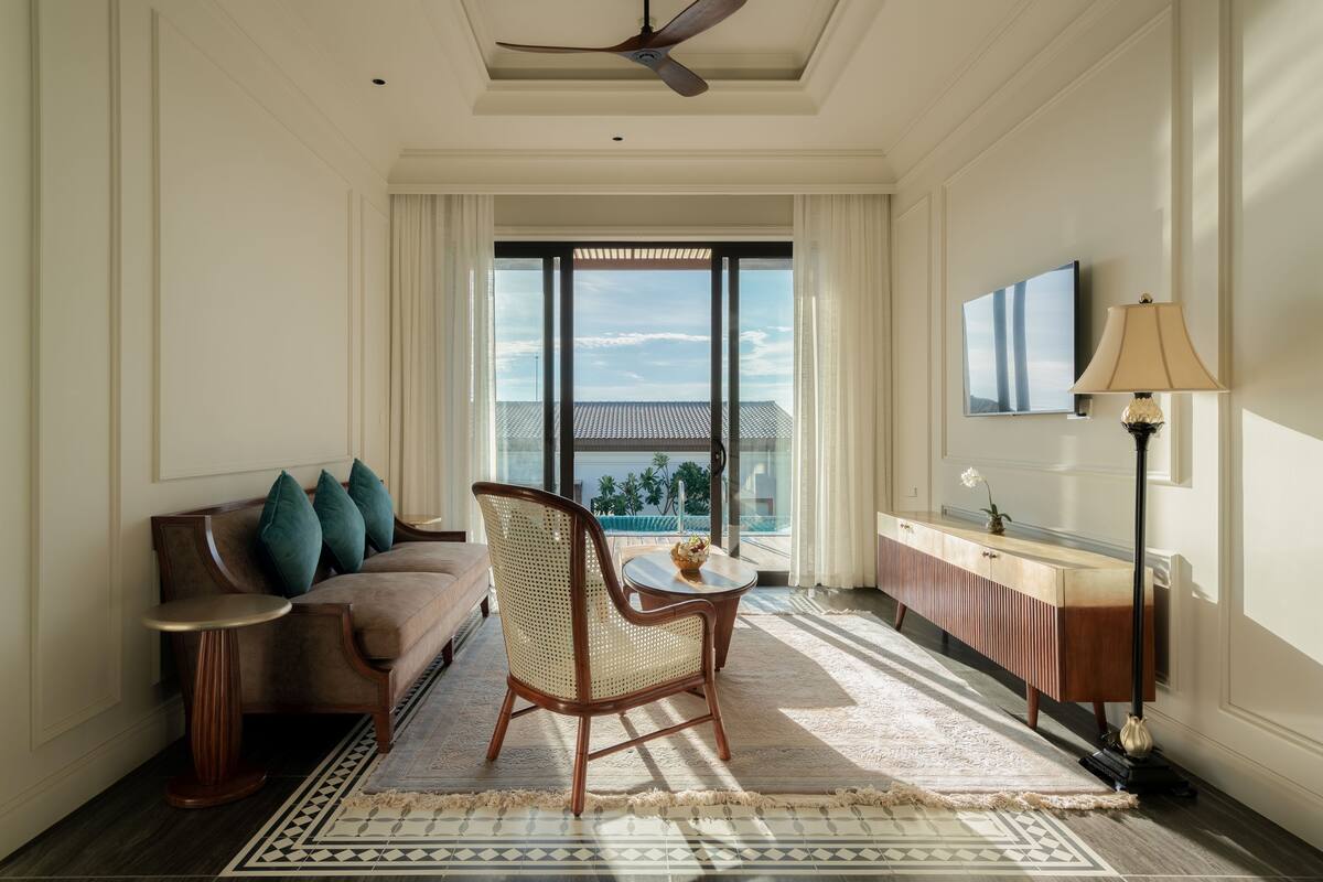 A hotel room with a sofa, a chair, a small table, a lamp, a TV, and a view of the outside.