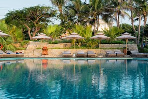 A swimming pool with lounge chairs and umbrellas, surrounded by palm trees and a stone wall.