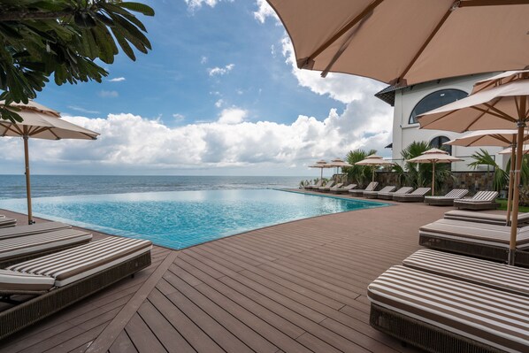 A poolside area with lounge chairs and umbrellas, overlooking a clear blue pool and ocean.