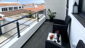 Un balcón con vistas a la ciudad, un sofá de mimbre negro, una mesita con una botella y un vaso, y una planta en maceta.
