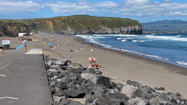 Una playa con costa rocosa, gente tomando el sol y nadando, y un cielo despejado.