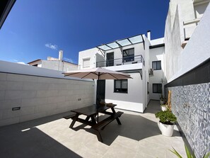 Un patio moderno al aire libre con una mesa y sillas, un nivel superior con paredes de vidrio y plantas en macetas.