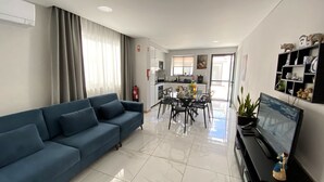 Una sala moderna con un sofá azul, una mesa de comedor con sillas, un televisor de pantalla plana y una cocina al fondo.