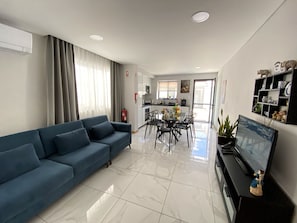 A modern living room with a blue sofa, a dining table with chairs, a flat-screen TV, and a kitchen in the background.