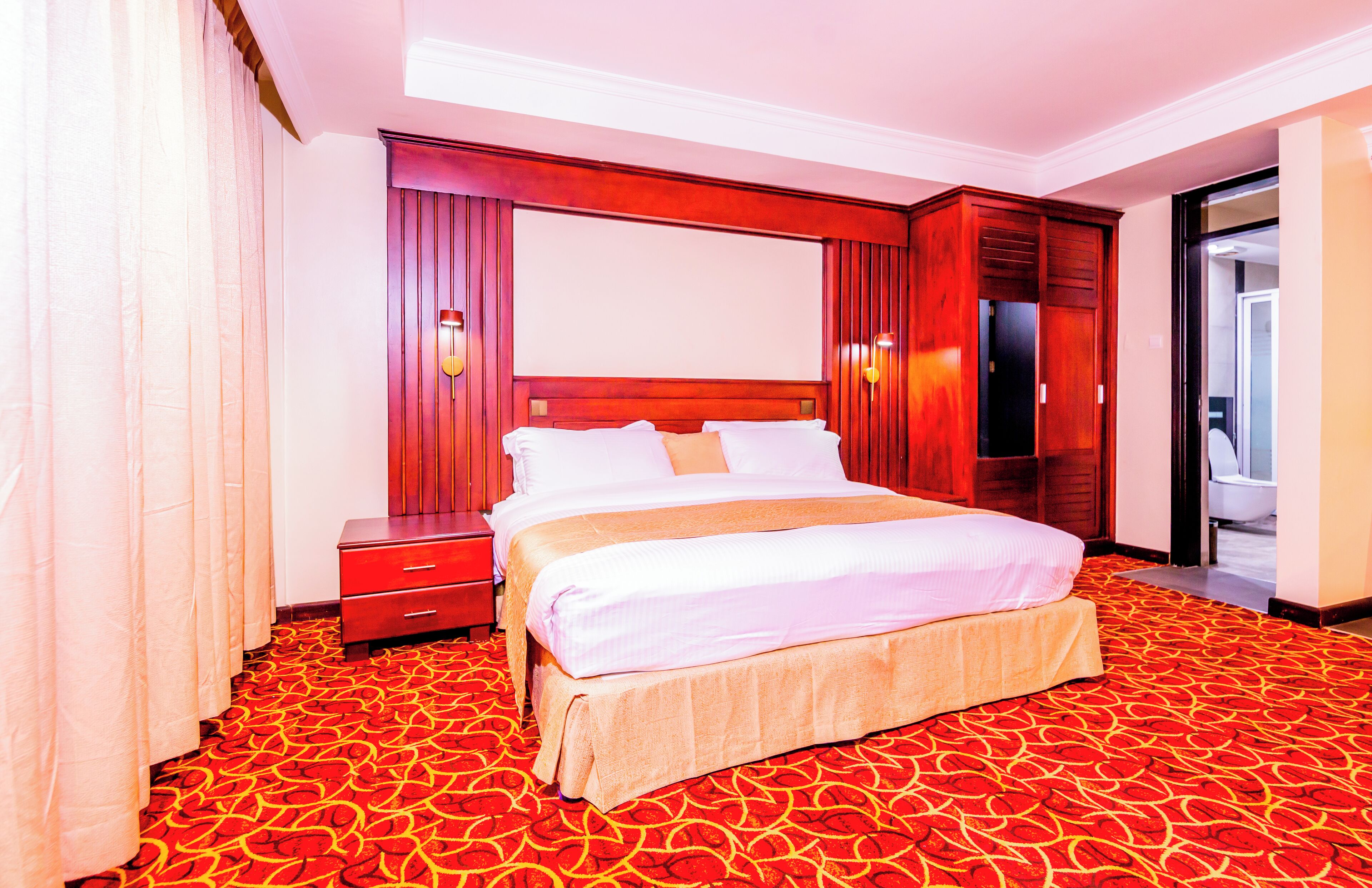 Une chambre d’hôtel avec un grand lit, une tête de lit en bois, une table de chevet, une télévision et une salle de bain visible à travers une porte ouverte.