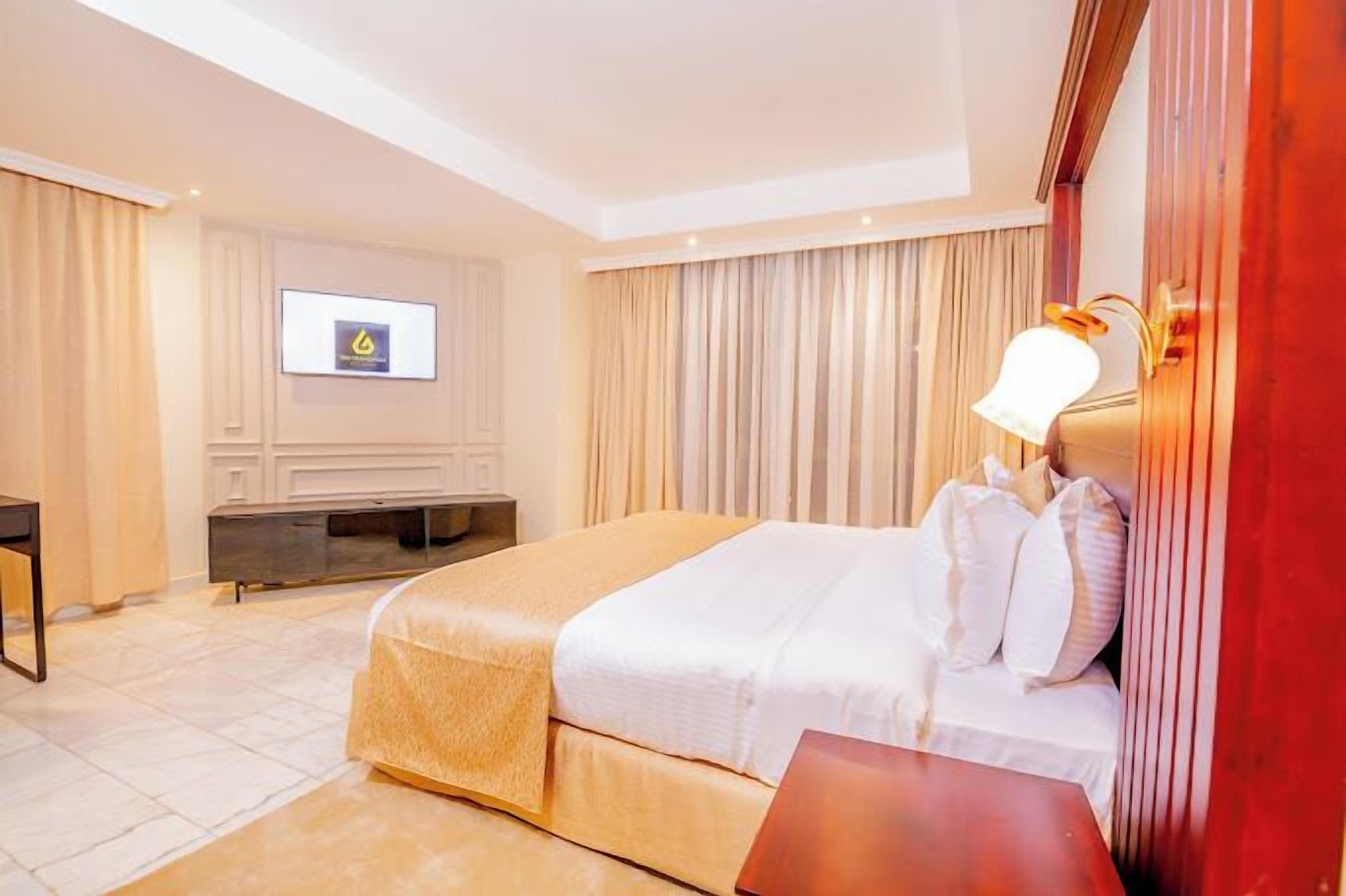A hotel room with a bed, a TV mounted on the wall, a desk, and a lamp.