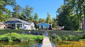 A wooden dock leading to a house with a covered patio and outdoor seating. - Loon Lodge - Super Cute, Lakefront, Bonfire Ring, and Private Dock! (Oscoda)