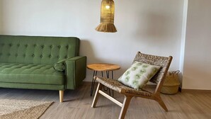 A green sofa, a wicker chair with a cushion, a small round table, and a hanging light fixture in a room with wooden flooring. - Apartamento Miramar (Denia)