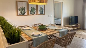 A dining area with a wooden table, wicker chairs, and a large pendant light. There are framed cactus paintings on the wall and a television in the corner. - Apartamento Miramar (Denia)
