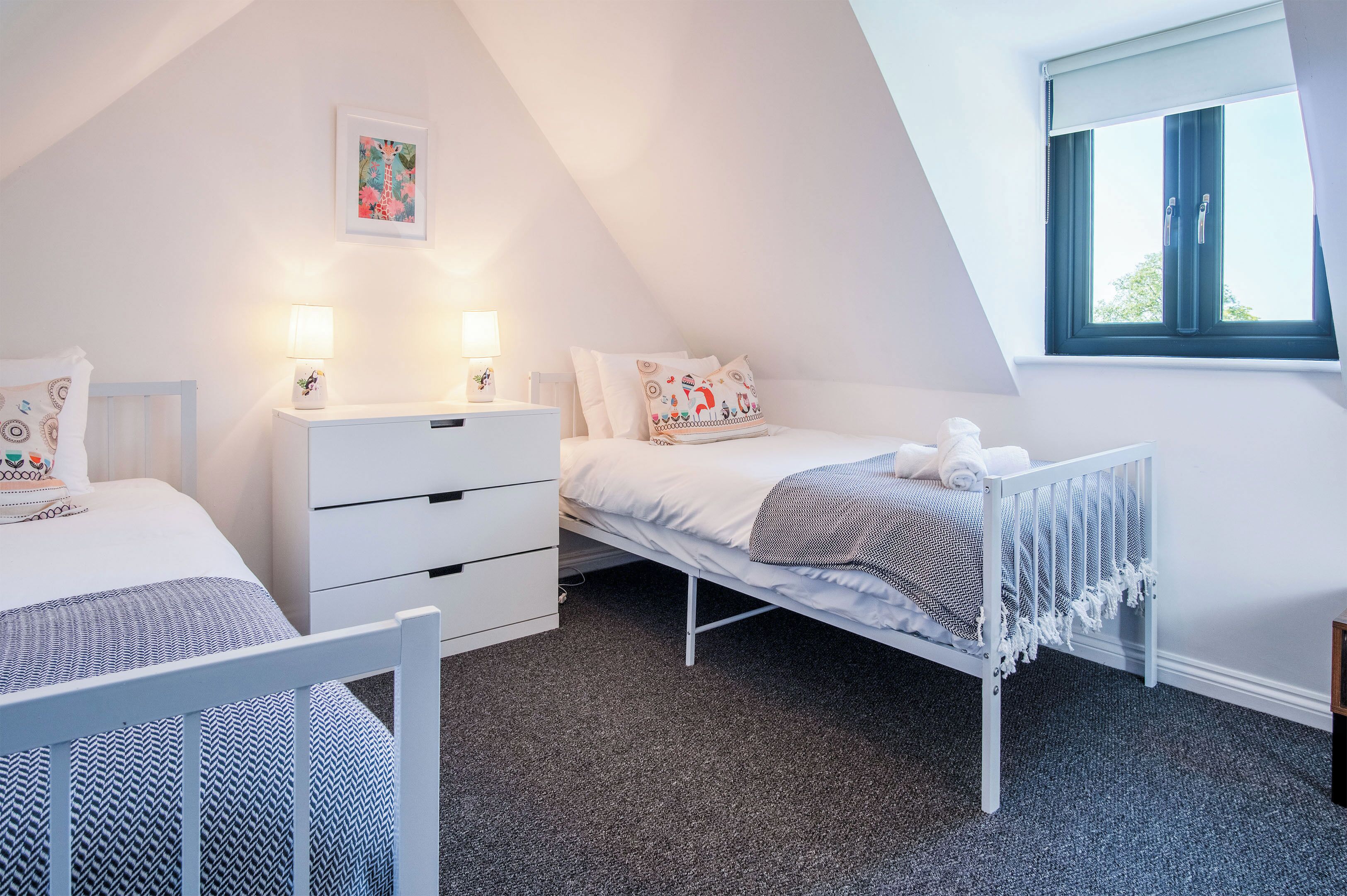 Ein Schlafzimmer mit schrägen Wänden, einem Bett mit weißer Bettwäsche, einem Nachttisch mit zwei Lampen und einem Fenster mit Blick ins Freie.