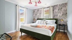 A bedroom with a green bed, white bedding, patterned wallpaper, a small side table, and a window with curtains. - The Islander | 2BR Riverland Terrace, Beach & DT (Charleston)