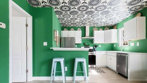 Una cucina con pareti verdi, mobili bianchi e un soffitto decorato. Sono presenti due sgabelli azzurri e un frigorifero.