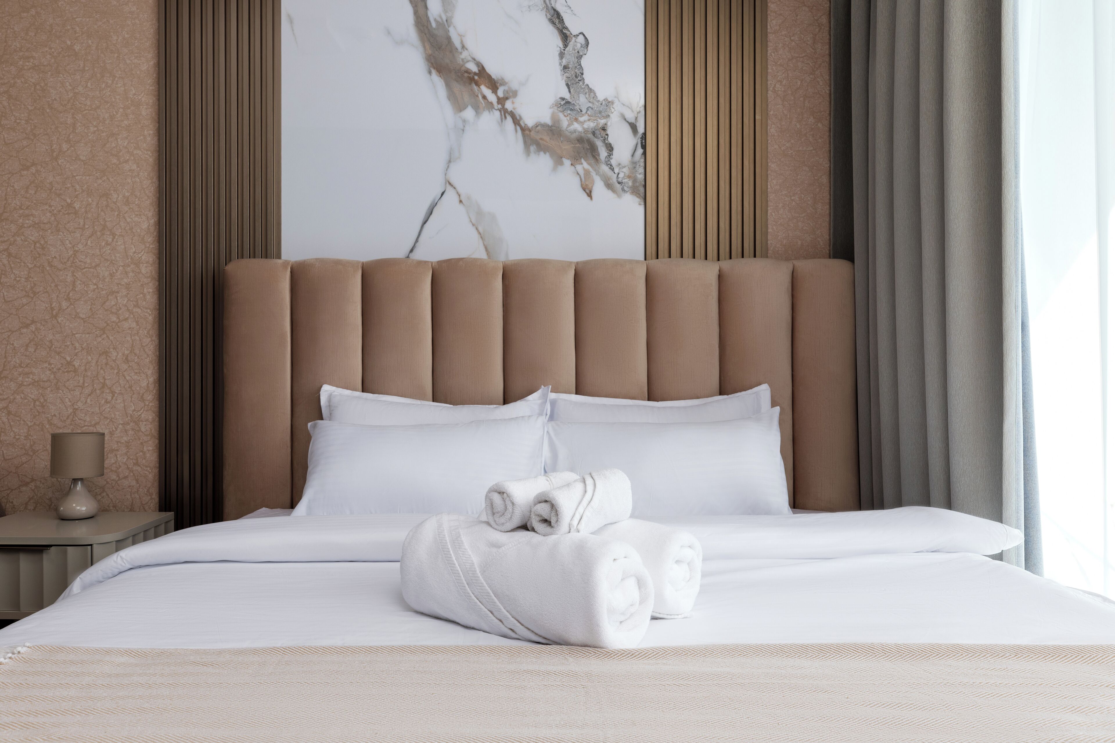 A neatly made bed with white linens and rolled towels, a headboard, and a nightstand with a lamp.