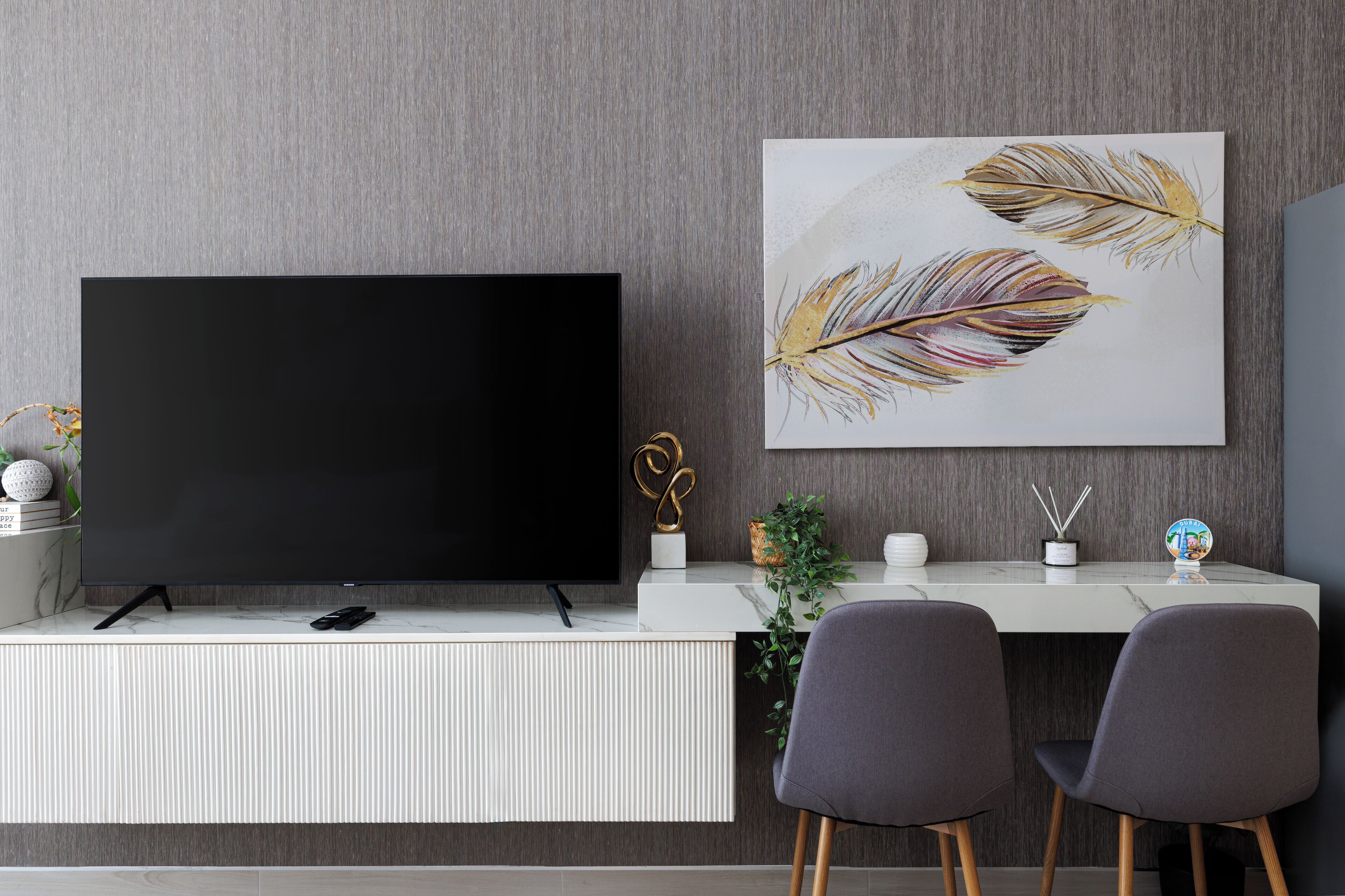 A modern living room with a large flat-screen TV, a white console table, two grey chairs, and a wall-mounted artwork of feathers.