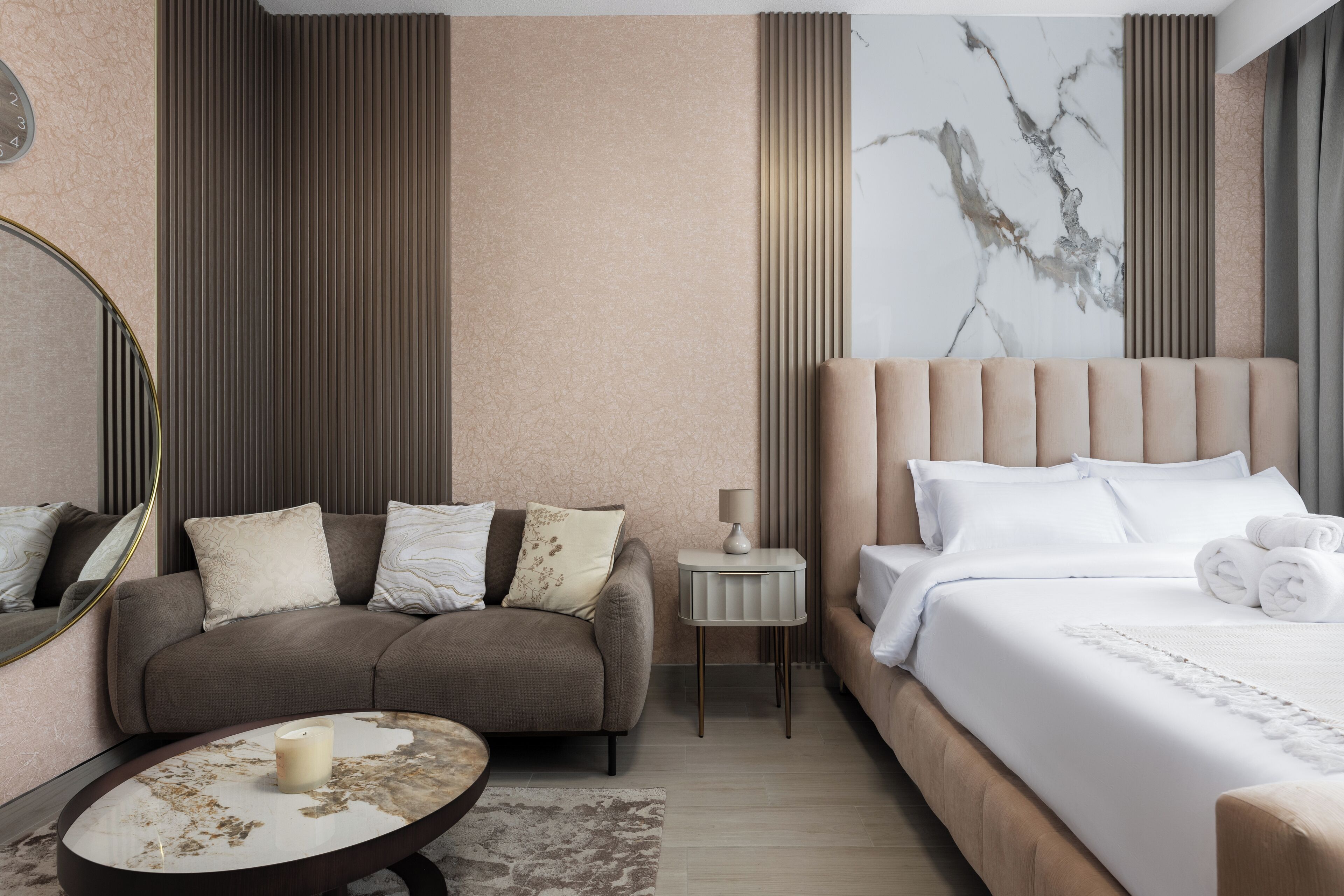 A modern hotel room with a sofa, a round coffee table, a bed with white linens, and a marble-patterned wall.