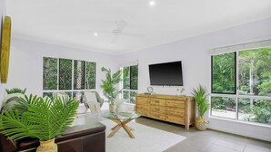 A modern living room with a glass coffee table, a brown sofa, a wooden cabinet, and large windows offering a view of greenery. - Palm Cove Studio Apartments (Palm Cove)