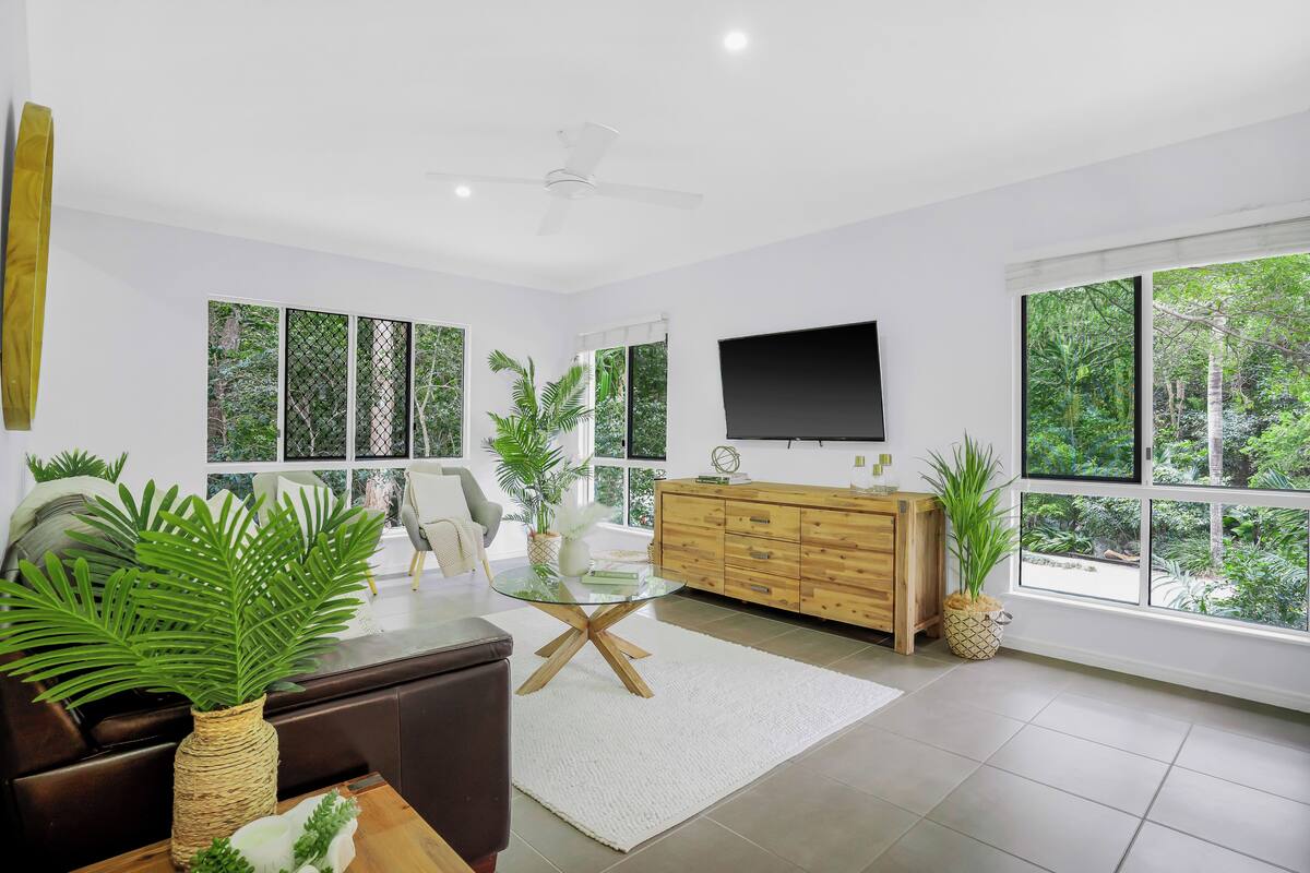 Ein modernes Wohnzimmer mit einem Glastisch, einem braunen Sofa, einem Holzkabinett und großen Fenstern mit Blick auf Grünflächen.