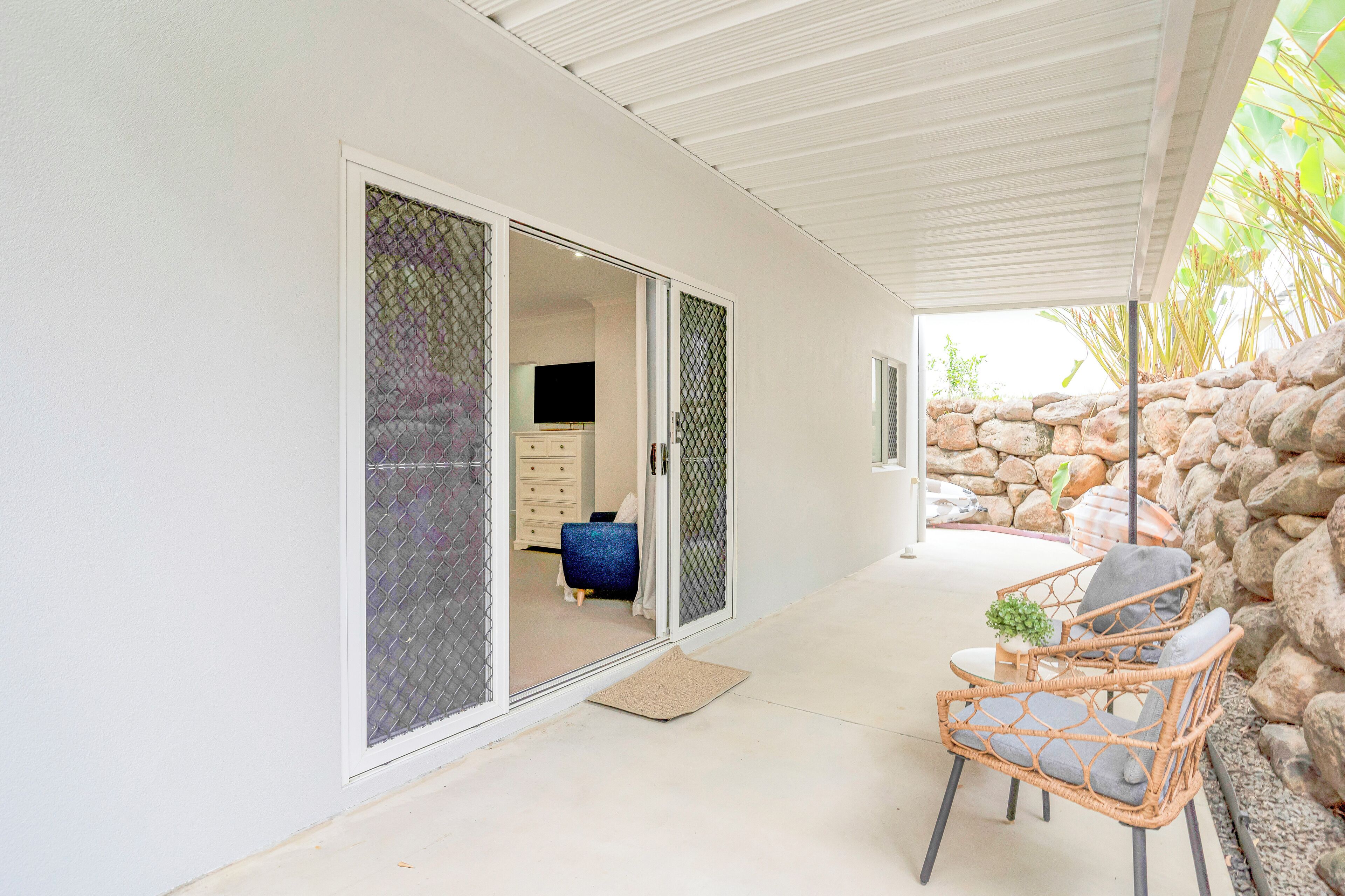 A covered patio with wicker chairs and a small table, a stone wall, and a sliding glass door leading to a room with a TV and a blue chair.
