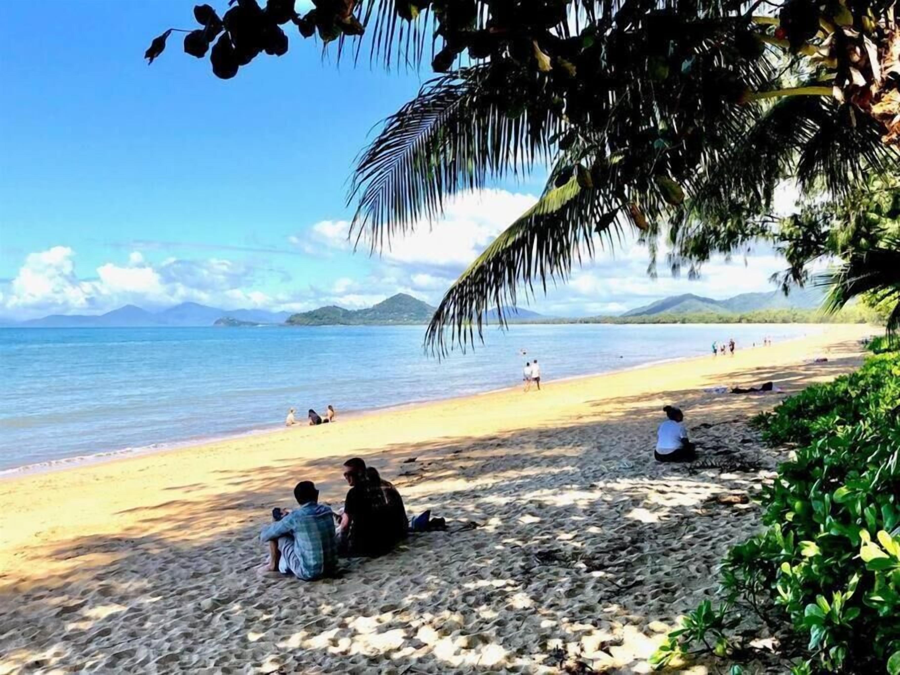 Ein Strand mit Menschen, die sitzen und spazieren gehen, Palmen und Bergen im Hintergrund.