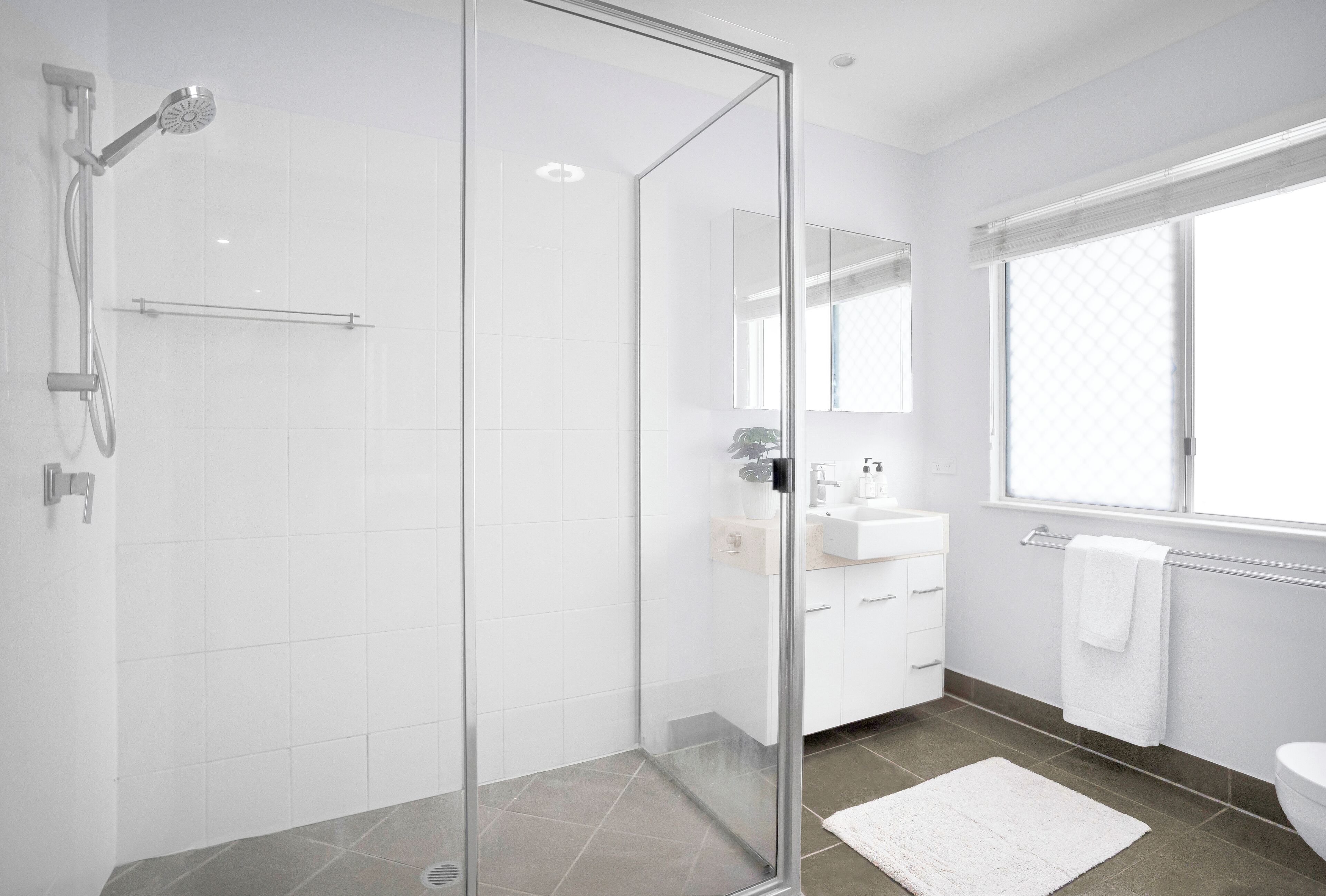 A modern bathroom with a glass shower enclosure, white tiles, a large mirror, and a window providing natural light.