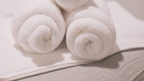 Three neatly rolled white towels on a bed with a plain wall in the background.