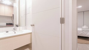 A modern hotel bathroom with a white sink, mirror, and a visible bed in the adjacent room.