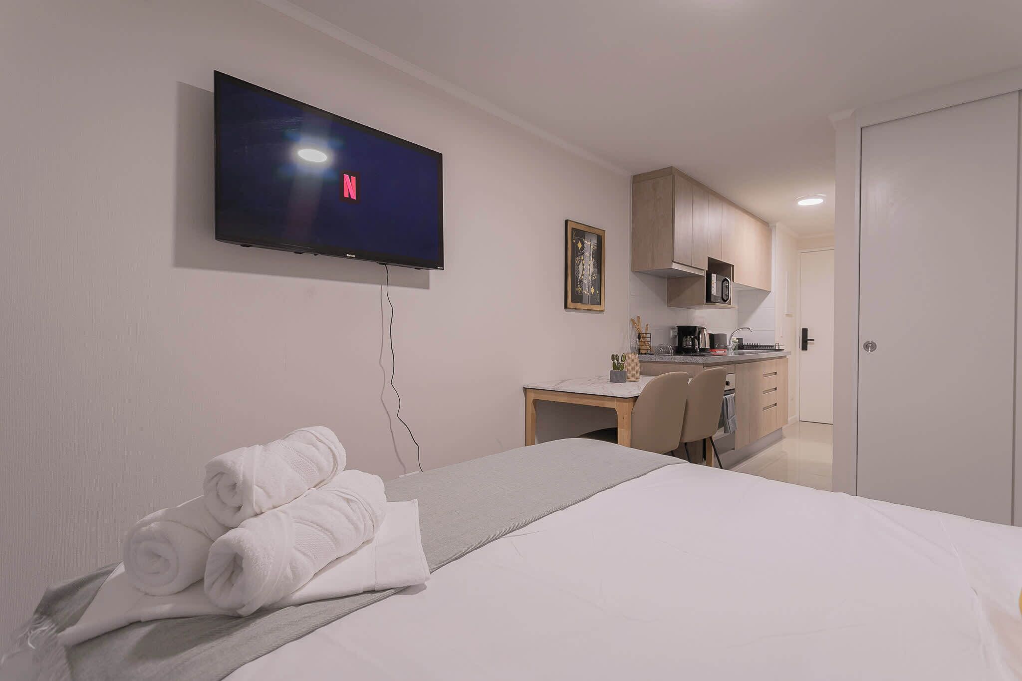 Habitación de hotel con cama, televisor montado en la pared, escritorio con silla y kitchenette.