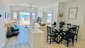 A spacious living area with a sofa, a dining table with chairs, a TV, and a balcony with a view of the ocean. - Hamilton Cove Villa 9 58 With Zero Steps (Avalon)