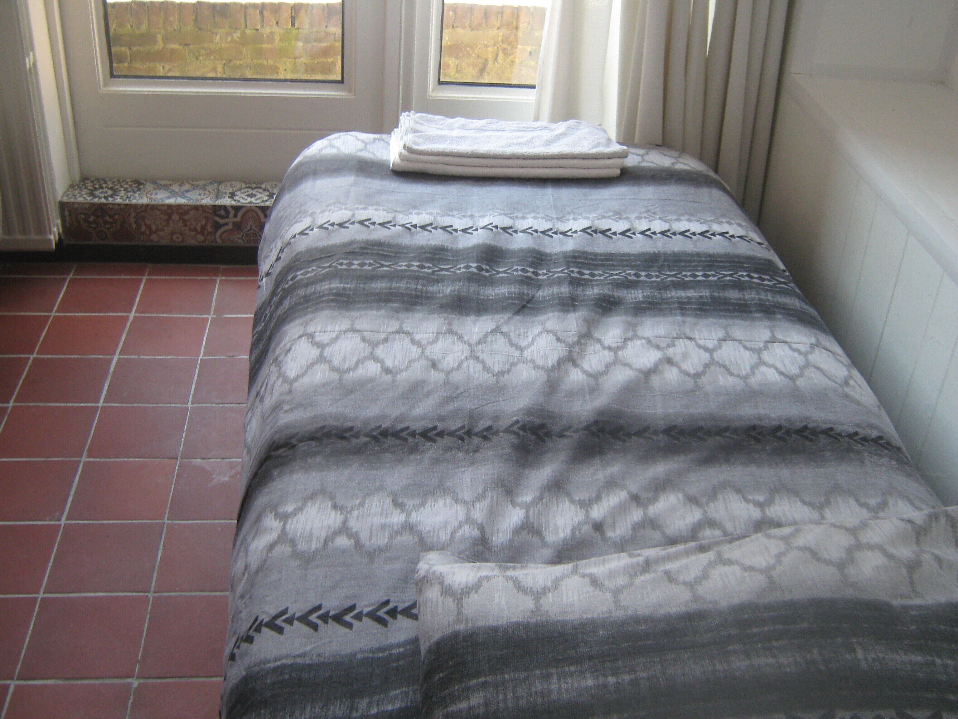 Una cama con una colcha estampada y toallas dobladas, un suelo de baldosas y una ventana con cortinas.