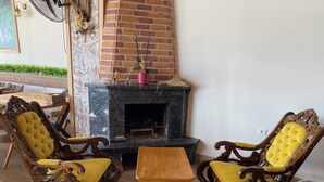 A cozy living room with a brick fireplace, two ornate wooden chairs with yellow cushions, and a small wooden table. - Bak cafe& pansiyon (Ayvalik)