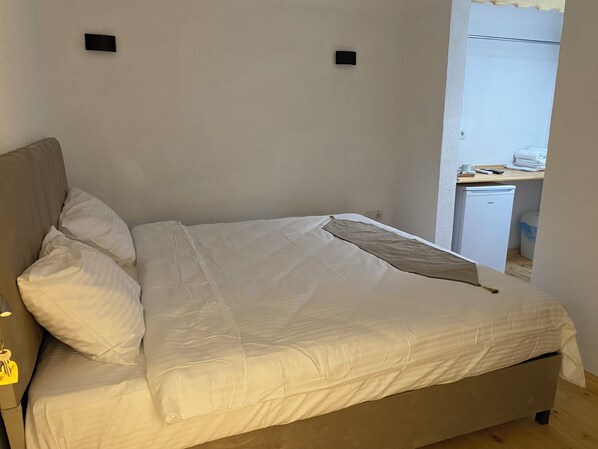 A neatly made bed with white bedding, a small bedside table with a lamp, and a wall-mounted TV. - Bak cafe& pansiyon (Ayvalik)