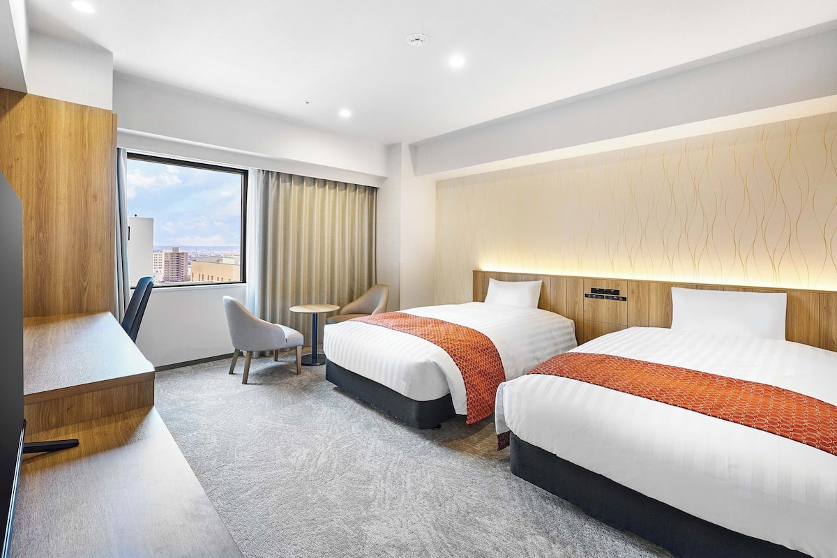 Ein Hotelzimmer mit zwei Betten, einem Schreibtisch, einem Sessel und Blick auf die Stadt.