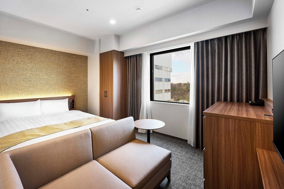 Ein modernes Hotelzimmer mit Bett, Sofa, Fernseher und einem Fenster mit Vorhängen.