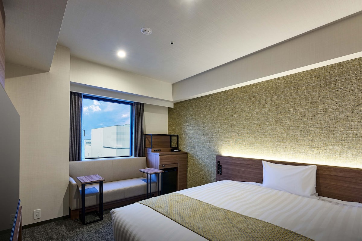 Ein modernes Hotelzimmer mit einem großen Bett, einem kleinen Schreibtisch, einem Fenster mit Vorhängen und einer strukturierten Wand.