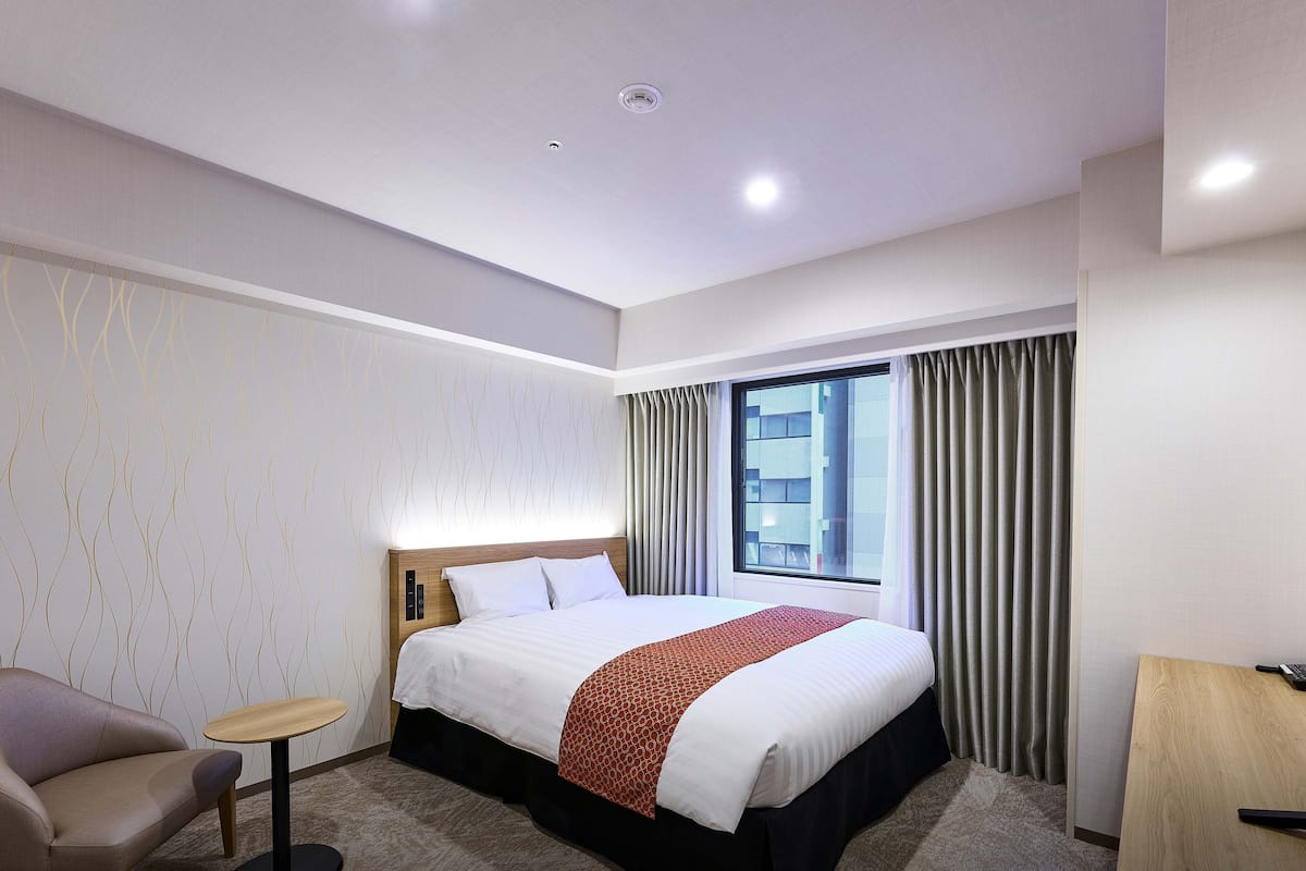 Ein Hotelzimmer mit Bett, einem kleinen Tisch, einem Sessel, einem Fernseher und einem Fenster mit Vorhängen.