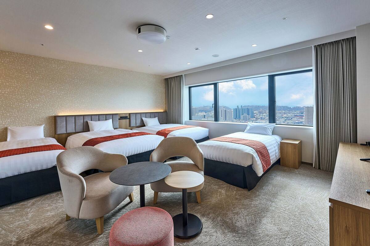 Ein Hotelzimmer mit zwei Betten, einem Schreibtisch, einem Sessel und Blick auf die Stadt.