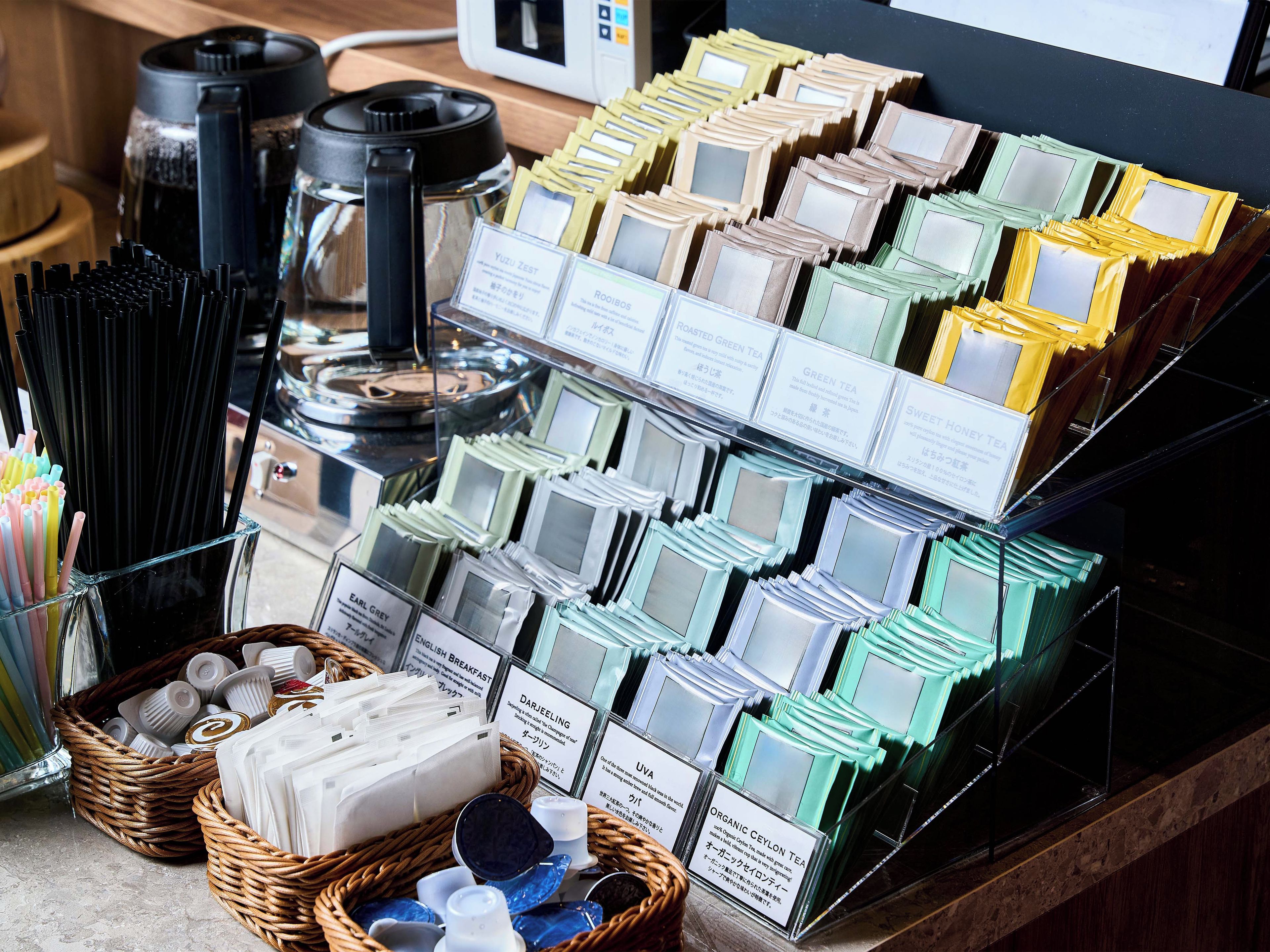 A display of tea varieties with labels, a coffee maker, and a basket of items on a counter.