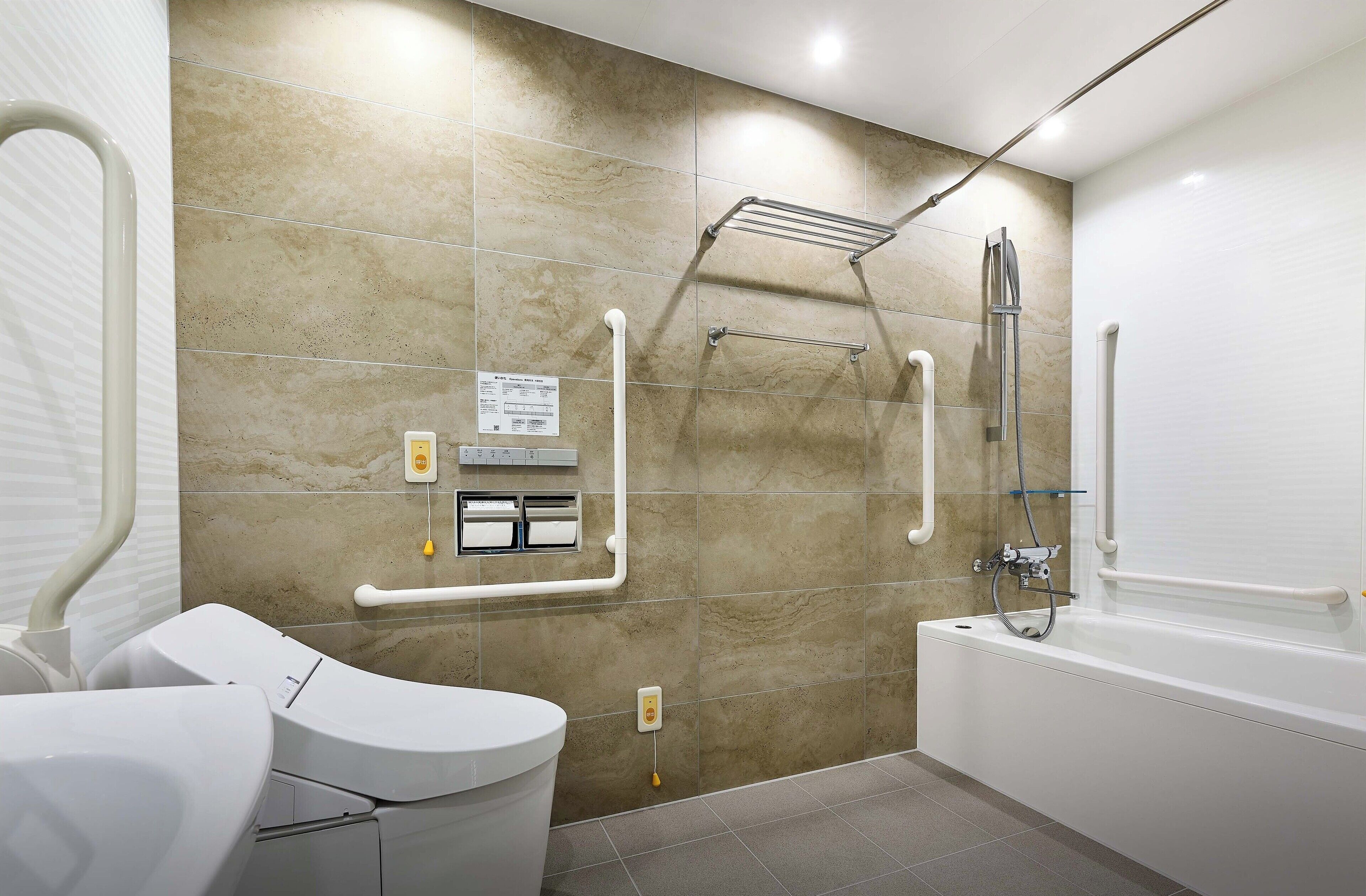 Une salle de bain moderne équipée d’une baignoire, de toilettes et d’un lavabo. Elle comprend également des barres d’appui et un siège de douche pour faciliter l’accès aux personnes à mobilité réduite.